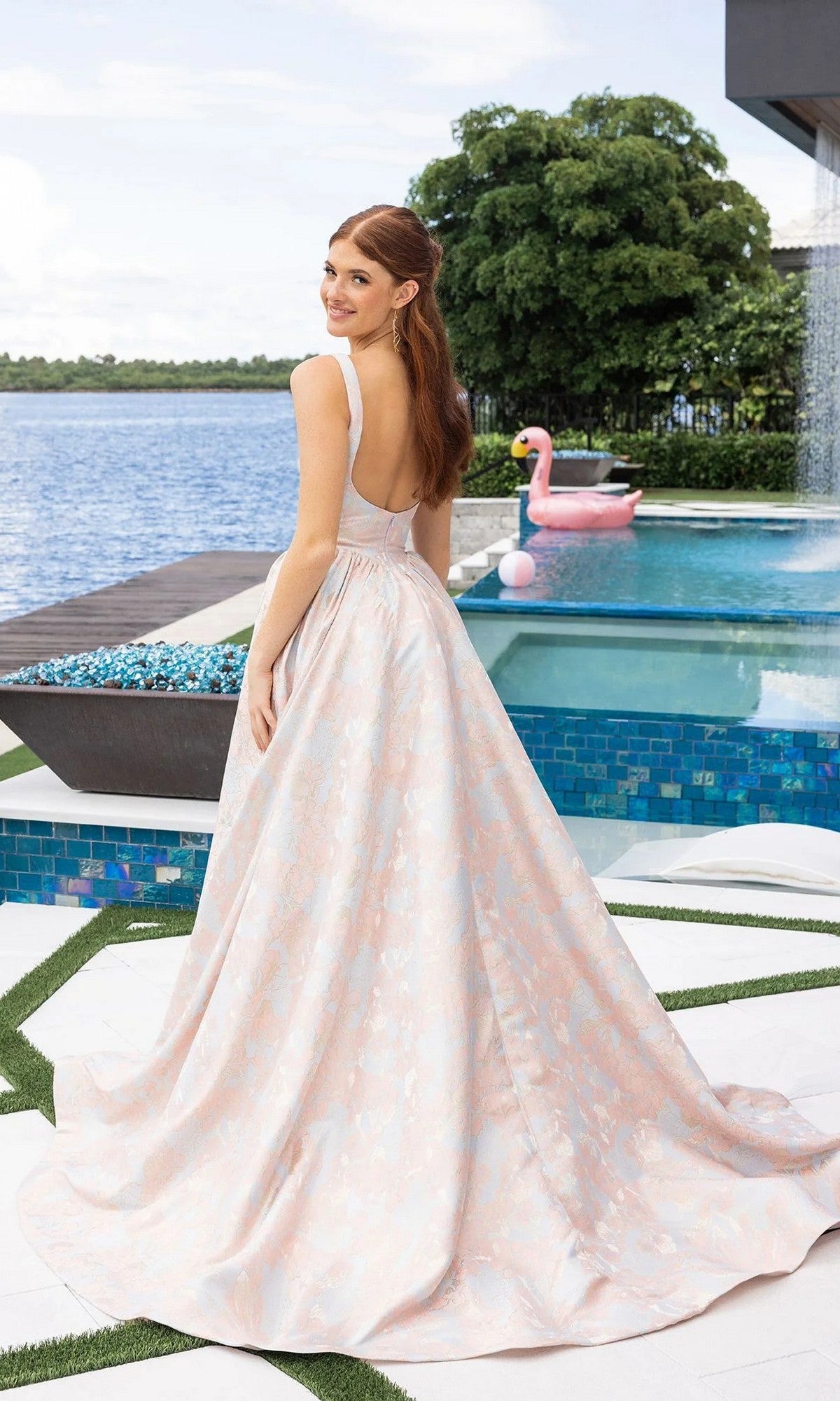 A woman in the Portia and Scarlett XO185 long formal dress, featuring pale pink and white jacquard, stands by a pool with a pink flamingo float and trees in the background, looking over her shoulder.